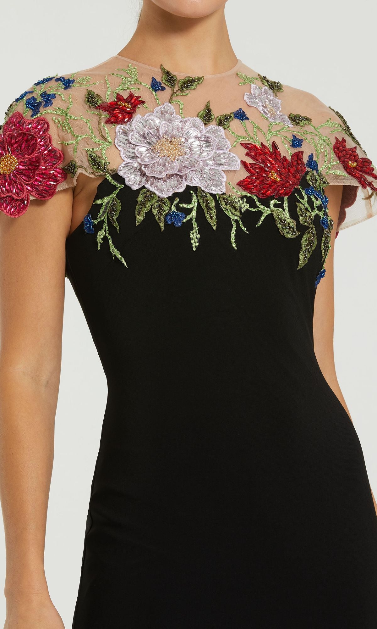 A woman wears the Mac Duggal 11817 Floral Long Black Formal Dress with a sheer neckline, embroidered colorful flowers, and green leaves.